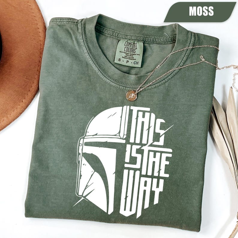 May include: A moss green t-shirt with a white graphic of a helmet and the text "THIS IS THE WAY." The shirt is made of a soft material and is styled with a gold necklace. The shirt is a small size.