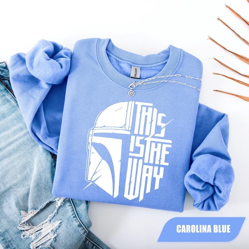 May include: Carolina blue sweatshirt with the text "THIS IS THE WAY" in white, along with a graphic of a helmet. A silver necklace is draped across the sweatshirt. The sweatshirt is displayed with a pair of blue jeans.