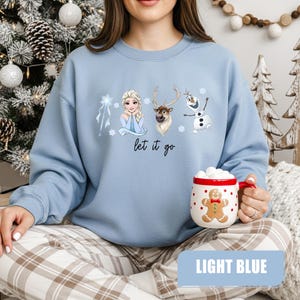 May include: Light blue sweatshirt featuring Elsa, a reindeer, and Olaf from Frozen, with the phrase "let it go." A gingerbread man mug with a red rim is held in the hand. The bottom of the image has a light blue rectangle with the words "LIGHT BLUE."
