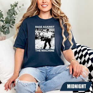 May include: Navy blue t-shirt featuring a black and white image with the text "RAGE AGAINST THE MACHINE". The graphic depicts police officers and a person. The word "MIDNIGHT" is at the bottom of the image.