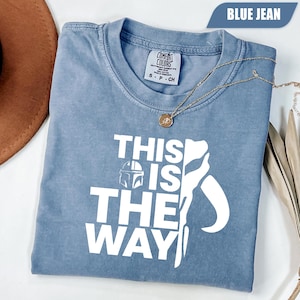 May include: A blue jean colored short-sleeve t-shirt with a white graphic print that reads THIS IS THE WAY, featuring a helmet icon and a stylized silhouette. The shirt has a Comfort Colors brand label at the inner neckline and a crew neck collar.