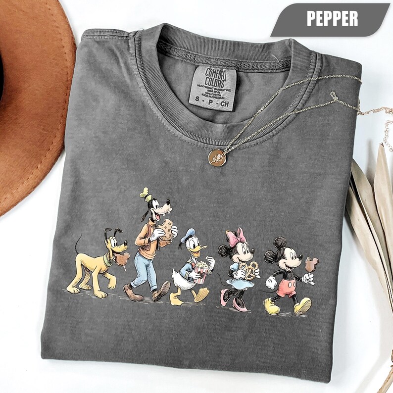 May include: A charcoal gray short-sleeve t-shirt with a crew neckline and a graphic print featuring Pluto, Goofy, Donald Duck, Minnie Mouse, and Mickey Mouse eating snacks. The inner neck label reads Comfort Colors, and the color is labeled as Pepper.