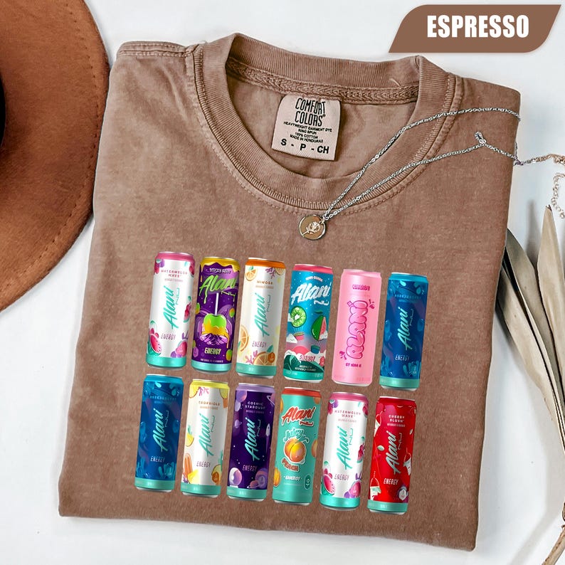May include: A brown crewneck t-shirt featuring a graphic print of twelve colorful Alani energy drink cans arranged in two rows. The shirt has a Comfort Colors brand label at the inner neckline and a silver-toned chain necklace resting on top.