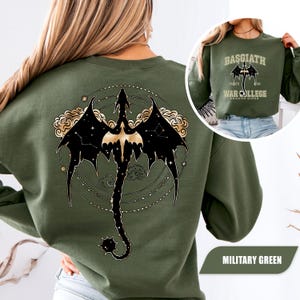 May include: Military green sweatshirt featuring a black dragon design with gold accents on the back. The dragon is set against a constellation and cloud background. The front of the sweatshirt has the text "BASGIATH WAR COLLEGE DRAGON RIDER".