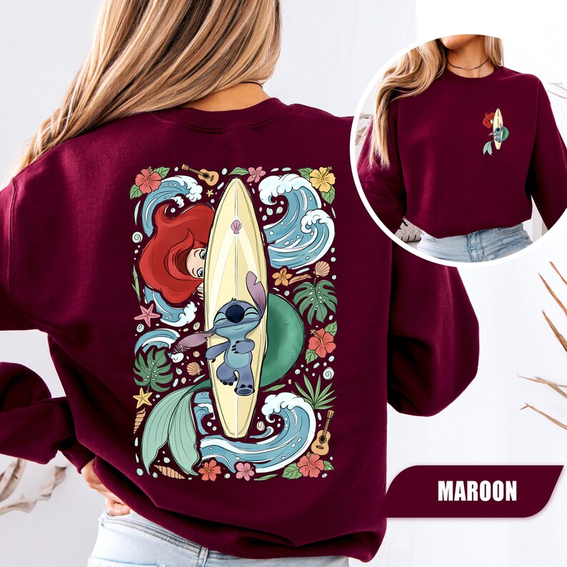 May include: A maroon crewneck sweatshirt featuring a large graphic on the back and a smaller version on the front left chest. The design depicts a blue alien character and a mermaid with red hair behind a surfboard, surrounded by waves and flowers.
