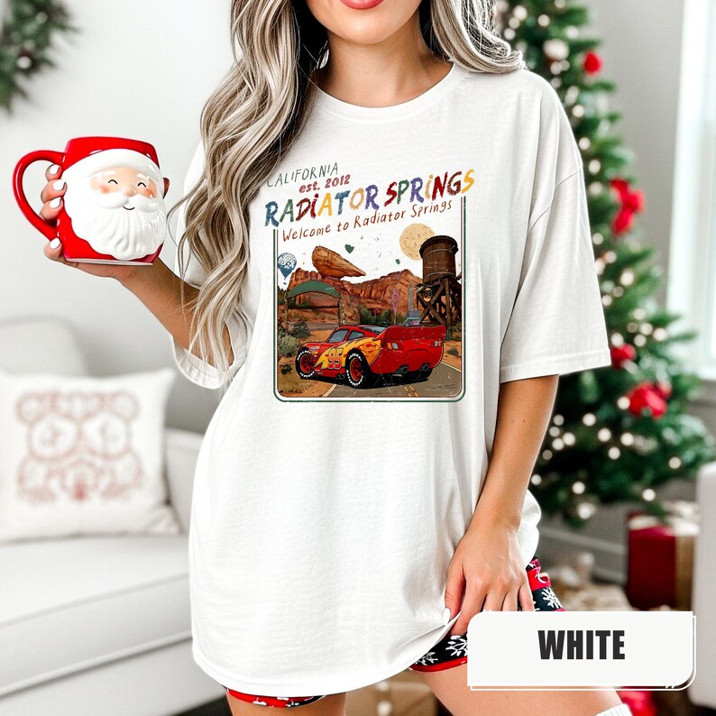 May include: A white t-shirt featuring a graphic print of a red race car in a desert landscape with the text "CALIFORNIA est. 2012 RADIATOR SPRINGS Welcome to Radiator Springs". The shirt is paired with a red Santa Claus shaped ceramic mug.
