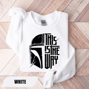 May include: White sweatshirt featuring a black graphic of a helmet and the text "THIS IS THE WAY." The sweatshirt is made of a soft material and has a crew neck. The word "WHITE" is displayed at the bottom.