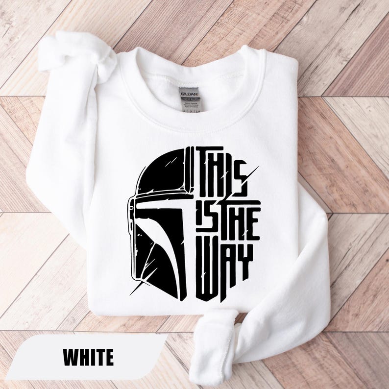 May include: White sweatshirt featuring a black graphic of a helmet and the text "THIS IS THE WAY." The sweatshirt is made of a soft material and has a crew neck. The word "WHITE" is displayed at the bottom.