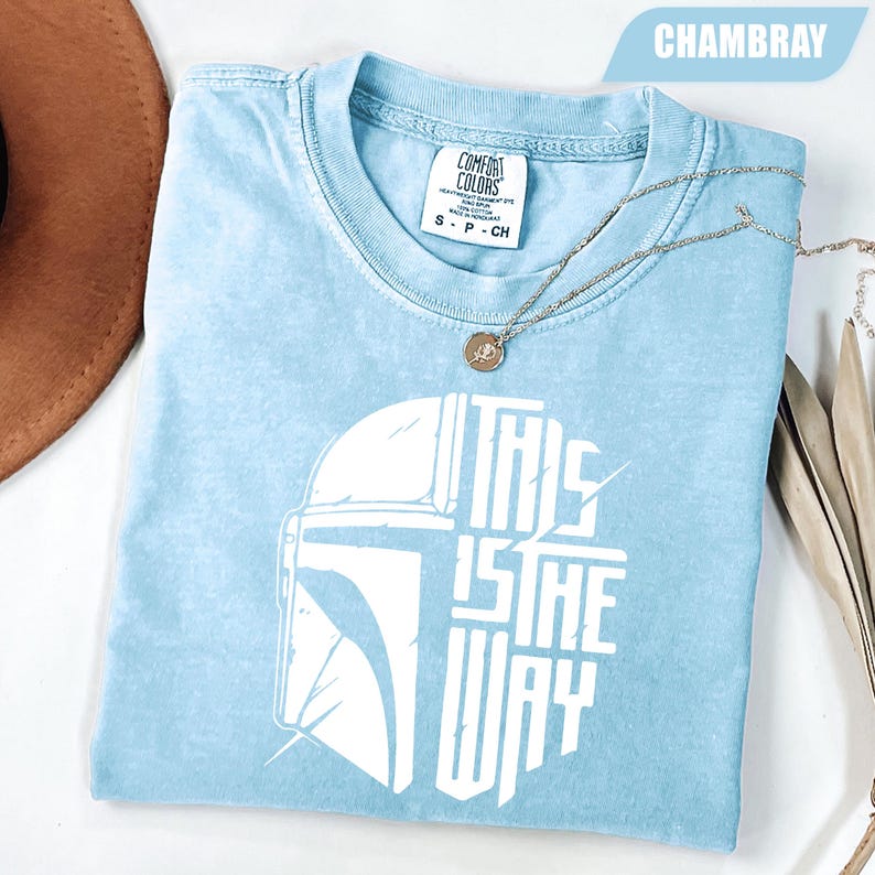 May include: Light blue t-shirt with a white graphic of a helmet and the text "THIS IS THE WAY." The shirt is made of Comfort Colors fabric and has a small tag at the neckline. A silver necklace with a pendant is draped across the shirt.