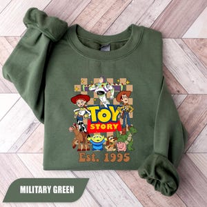 Puede incluir: Sudadera verde militar con un gr&aacute;fico de Toy Story con Woody, Buzz Lightyear y Jessie. El dise&ntilde;o incluye el texto "TOY STORY" y "Est. 1995". La sudadera est&aacute; hecha de un material suave.
