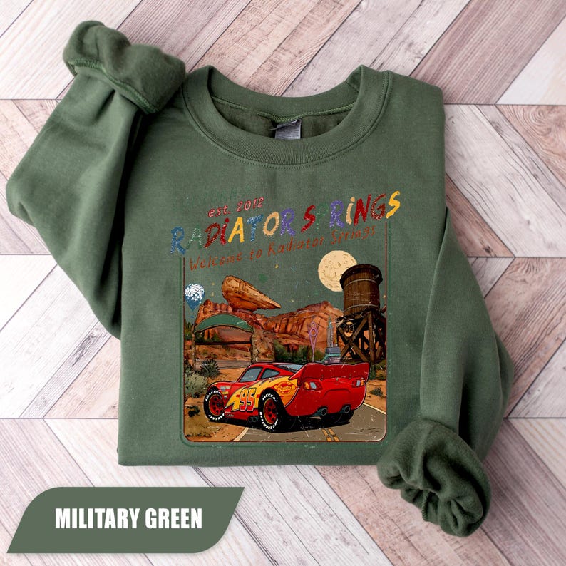 May include: A military green sweatshirt featuring a graphic print of a red race car with the number 95. The design includes the text "est. 2012 RADIATOR SPRINGS" and "Welcome to Radiator Springs" above a desert landscape with a water tower and moon.