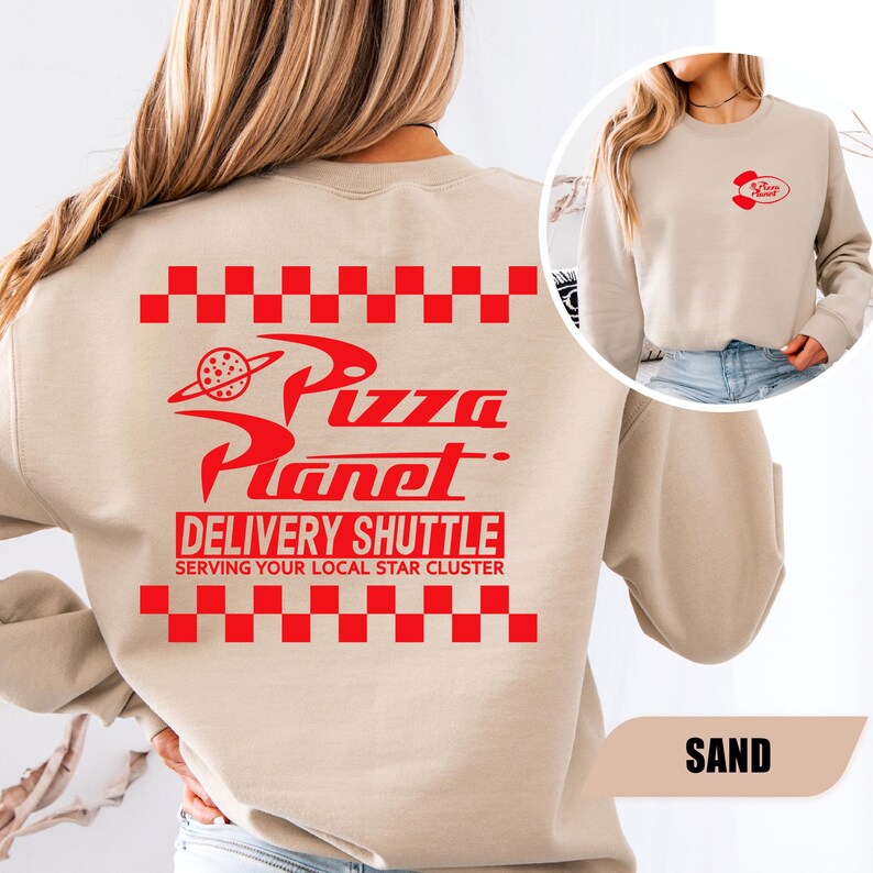May include: A sand-colored crewneck sweatshirt featuring a red graphic on the back with a checkered border, the text Pizza Planet, and the words DELIVERY SHUTTLE SERVING YOUR LOCAL STAR CLUSTER. A smaller red Pizza Planet logo is on the front chest.