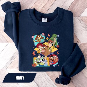 Puede incluir: Sudadera azul marino con un gr&aacute;fico colorido de personajes de dibujos animados. El dise&ntilde;o incluye un perro, un dinosaurio, un vaquero y otros personajes. La palabra "NAVY" est&aacute; impresa en la parte inferior.