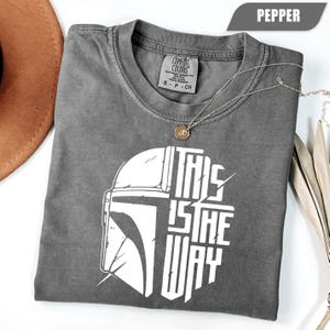 May include: Pepper-colored t-shirt with a white graphic of a helmet and the text "THIS IS THE WAY." The shirt is made of Comfort Colors heavyweight garment dye material. A silver necklace with a pendant is draped across the shirt.