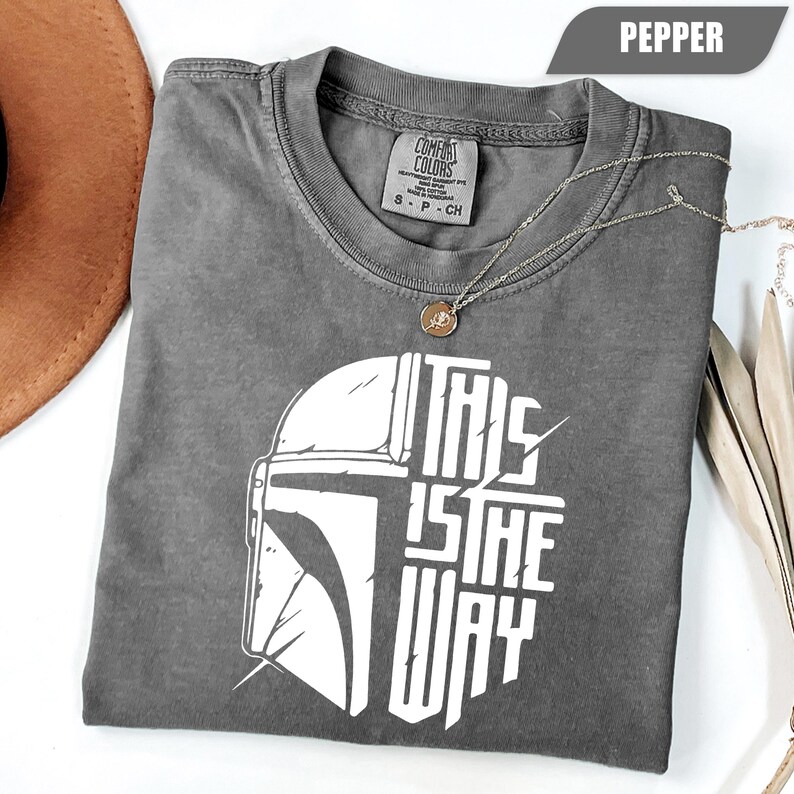 May include: Pepper-colored t-shirt with a white graphic of a helmet and the text "THIS IS THE WAY." The shirt is made of Comfort Colors heavyweight garment dye material. A silver necklace with a pendant is draped across the shirt.