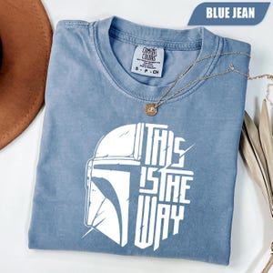 May include: Blue jean colored t-shirt with a white graphic of a helmet and the text "THIS IS THE WAY". The shirt has a crew neck and short sleeves. A gold necklace with a pendant is draped across the shirt.
