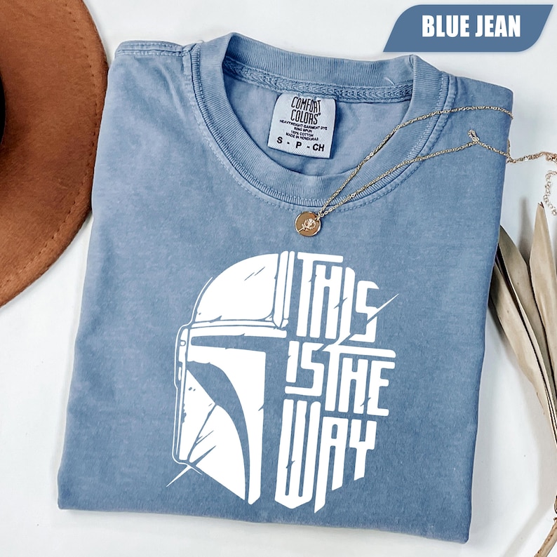 May include: Blue jean colored t-shirt with a white graphic of a helmet and the text "THIS IS THE WAY". The shirt has a crew neck and short sleeves. A gold necklace with a pendant is draped across the shirt.