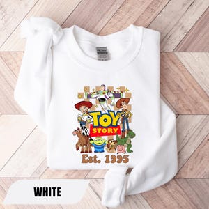 Puede incluir: Sudadera blanca con un estampado colorido de los personajes de Toy Story, incluyendo a Woody, Buzz Lightyear y Jessie. Las palabras "TOY STORY" est&aacute;n en letras grandes y amarillas, con "Est. 1995" debajo. La palabra "WHITE" est&aacute; en la parte inferior.