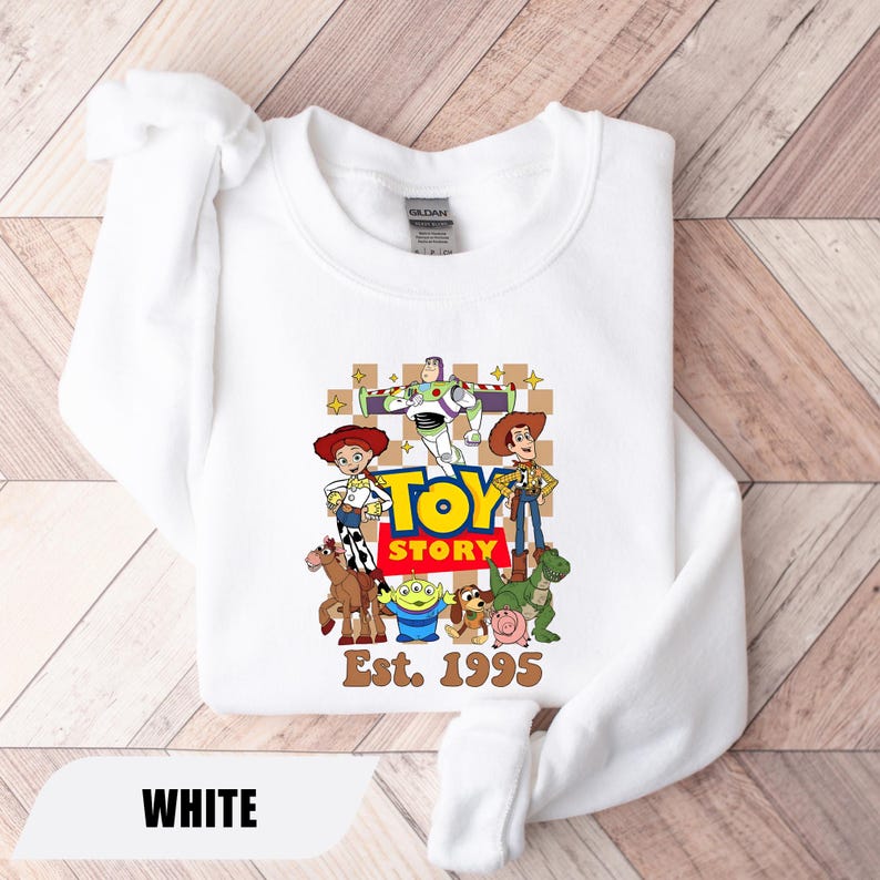 Puede incluir: Sudadera blanca con un estampado colorido de los personajes de Toy Story, incluyendo a Woody, Buzz Lightyear y Jessie. Las palabras "TOY STORY" est&aacute;n en letras grandes y amarillas, con "Est. 1995" debajo. La palabra "WHITE" est&aacute; en la parte inferior.