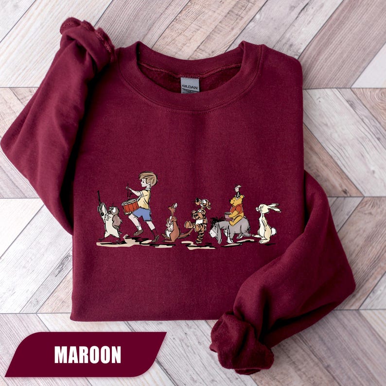 May include: Maroon sweatshirt featuring a line of cartoon characters, including a boy with a drum, a kangaroo, and a tiger. The word "MAROON" is displayed in a white font on a maroon banner at the bottom of the sweatshirt.