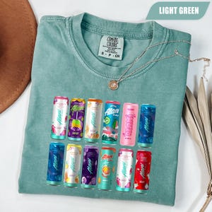 May include: A light green short-sleeve t-shirt featuring a graphic print of twelve colorful Alani energy drink cans arranged in two rows. The shirt has a crew neckline and a Comfort Colors brand label on the inside collar. A thin gold chain necklace rests on the fabric.