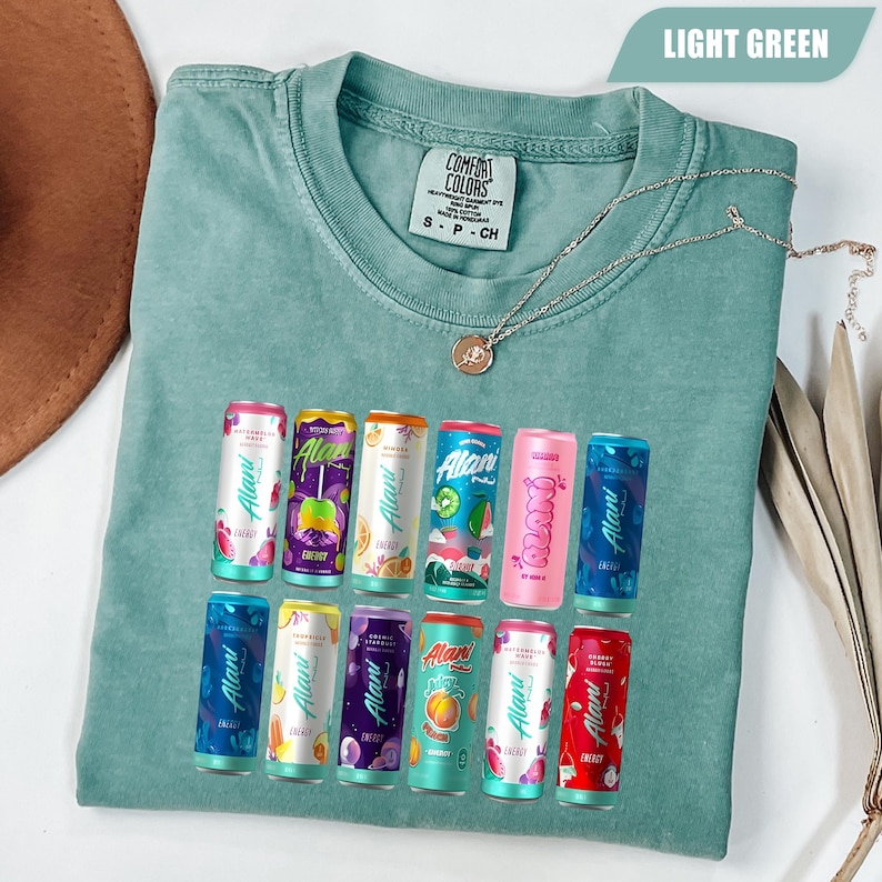 May include: A light green short-sleeve t-shirt featuring a graphic print of twelve colorful Alani energy drink cans arranged in two rows. The shirt has a crew neckline and a Comfort Colors brand label on the inside collar. A thin gold chain necklace rests on the fabric.