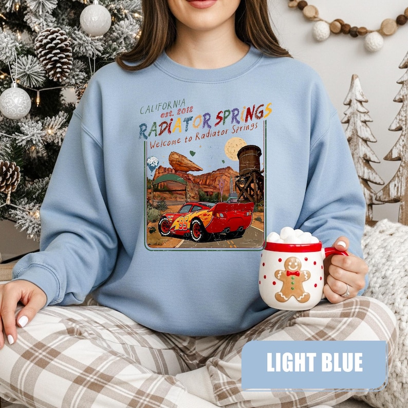 May include: A light blue crewneck sweatshirt featuring a graphic print of a red race car in a desert landscape with the text California est. 2012 Radiator Springs Welcome to Radiator Springs. A white mug with a gingerbread man design is also shown.