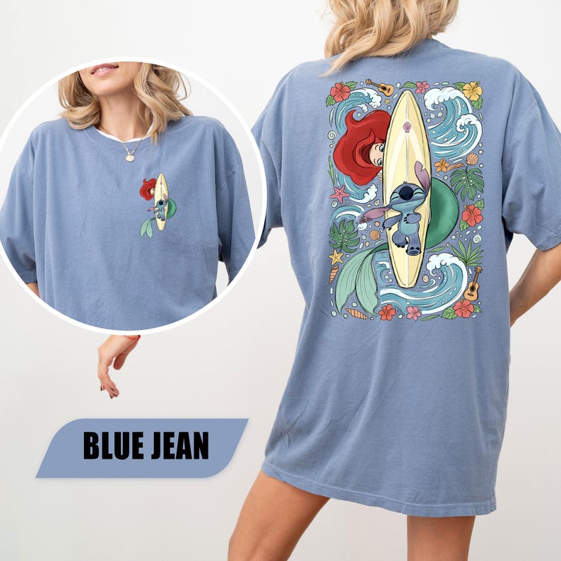 May include: A blue jean colored t-shirt featuring a small graphic on the front left chest and a large graphic on the back. Both graphics depict a blue alien character and a mermaid with red hair surfing on a surfboard surrounded by tropical flowers, leaves, waves, seashells, and a small ukulele.
