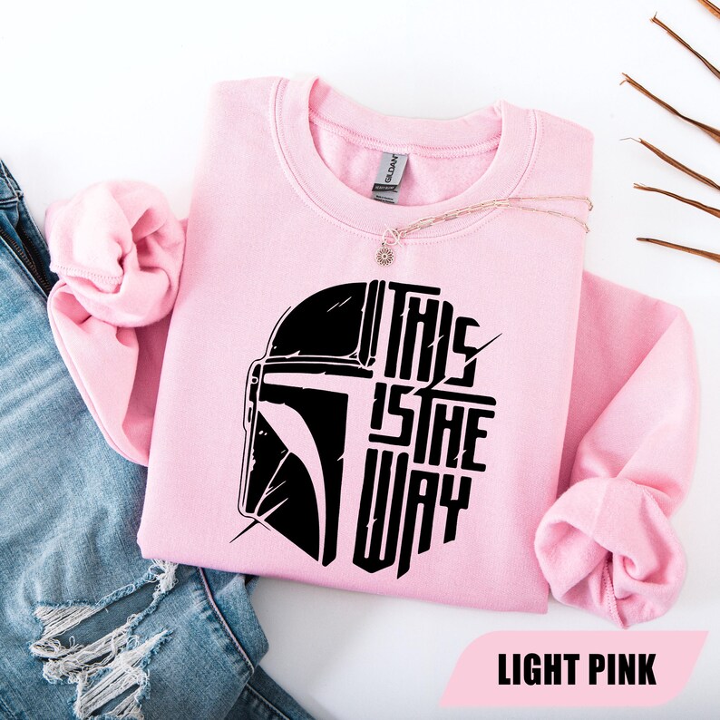 May include: Light pink sweatshirt with a black graphic of a helmet and the text "THIS IS THE WAY." The sweatshirt is a crew neck style and is made of a soft material. A gold necklace with a pendant is draped on the sweatshirt.