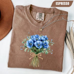 Może przedstawiać: Koszulka Comfort Colors w kolorze espresso z niebieskim wzorem bukietu kwiat&oacute;w. Bukiet jest związany sznurkową kokardą. Koszulka ma okrągły dekolt i kr&oacute;tkie rękawy. Srebrny naszyjnik z wisiorkiem jest zarzucony na koszulkę.