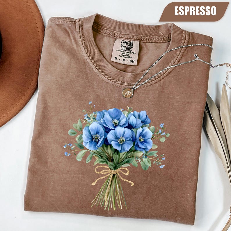 Może przedstawiać: Koszulka Comfort Colors w kolorze espresso z niebieskim wzorem bukietu kwiat&oacute;w. Bukiet jest związany sznurkową kokardą. Koszulka ma okrągły dekolt i kr&oacute;tkie rękawy. Srebrny naszyjnik z wisiorkiem jest zarzucony na koszulkę.
