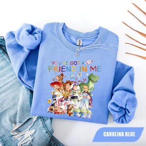 May include: A light blue crewneck sweatshirt featuring a colorful graphic print of various animated toy characters and the text "YOU'VE GOT A FRIEND IN ME" in multi-colored lettering. The garment has long sleeves and a ribbed collar, cuffs, and hem.