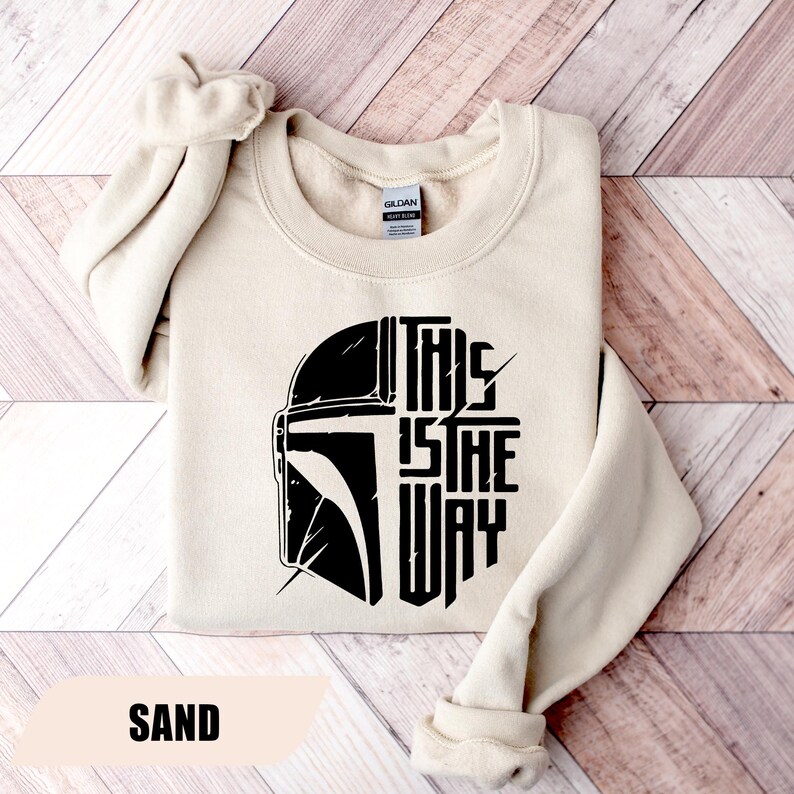 May include: Sand-colored sweatshirt featuring a black graphic of a helmet and the text "THIS IS THE WAY." The sweatshirt has a crew neck and long sleeves, with the sleeves cuffed. The word "SAND" is printed at the bottom.