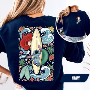 May include: A navy blue crewneck sweatshirt featuring a large graphic on the back and a smaller version on the front left chest. The design depicts a cartoon character with a surfboard, a mermaid, tropical flowers, waves, and a small guitar.
