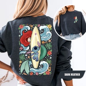 May include: A dark heather gray crewneck sweatshirt featuring a large back graphic of a blue alien character on a surfboard, a red-haired mermaid, tropical flowers, green leaves, ocean waves, and a small guitar. A small matching graphic is on the front.
