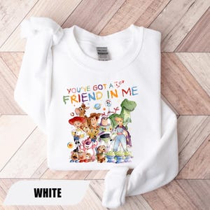 May include: A white crewneck sweatshirt featuring a colorful graphic print of various animated toy characters and the text "YOU'VE GOT A FRIEND IN ME" in a multi-colored, crayon-style font. The design includes characters like a cowboy, space ranger, and dinosaur.