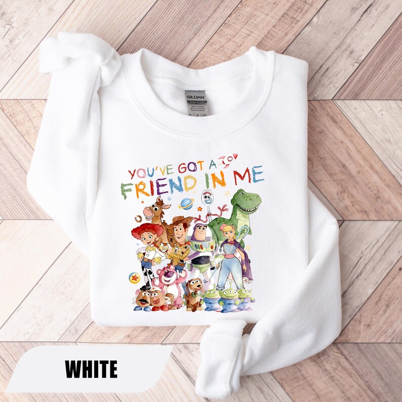 May include: A white crewneck sweatshirt featuring a colorful graphic print of various animated toy characters and the text "YOU'VE GOT A FRIEND IN ME" in a multi-colored, crayon-style font. The design includes characters like a cowboy, space ranger, and dinosaur.