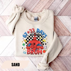 May include: Sand-colored sweatshirt featuring a graphic of Spider-Man with the text "67+33 DAYS BRUH". The design includes a checkered background, stars, and school-themed illustrations. The sweatshirt has a crew neck and long sleeves.