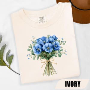 Może przedstawiać: Kremowa koszulka z akwarelowym bukietem niebieskich kwiat&oacute;w związanym sznurkiem. Etykieta marki Comfort Colors jest widoczna przy dekolcie. Słowo "IVORY" jest wydrukowane na dole obrazu.
