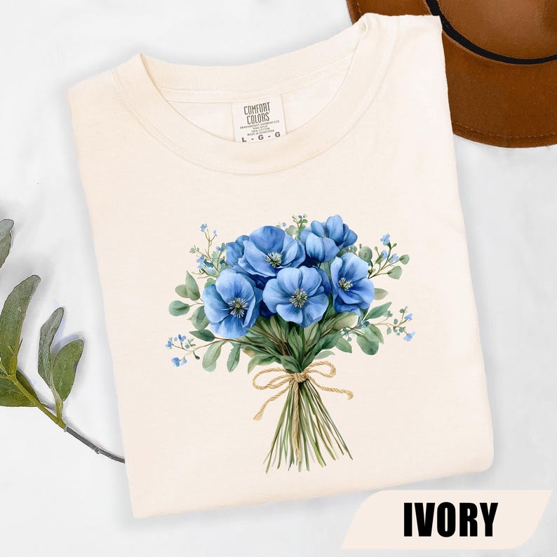 Może przedstawiać: Kremowa koszulka z akwarelowym bukietem niebieskich kwiat&oacute;w związanym sznurkiem. Etykieta marki Comfort Colors jest widoczna przy dekolcie. Słowo "IVORY" jest wydrukowane na dole obrazu.