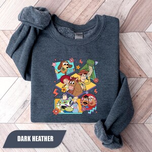 Puede incluir: Sudadera gris oscuro con un dise&ntilde;o gr&aacute;fico colorido de personajes de dibujos animados. El dise&ntilde;o incluye un perro, un dinosaurio, un vaquero, un guardabosques espacial y otros personajes. El texto "DARK HEATHER" est&aacute; en la parte inferior.