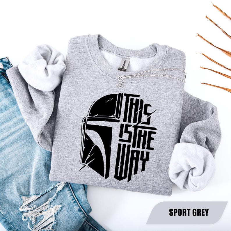 May include: Sport grey sweatshirt with a black graphic of a helmet and the text "THIS IS THE WAY". The sweatshirt has a crew neck and long sleeves with white lining. A silver necklace is draped across the top.