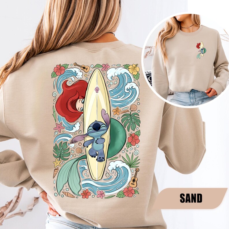 May include: A sand-colored crewneck sweatshirt featuring a large graphic on the back of a blue alien character on a surfboard, surrounded by a red-haired mermaid, ocean waves, tropical flowers, seashells, and ukuleles. A small matching graphic is on the front.