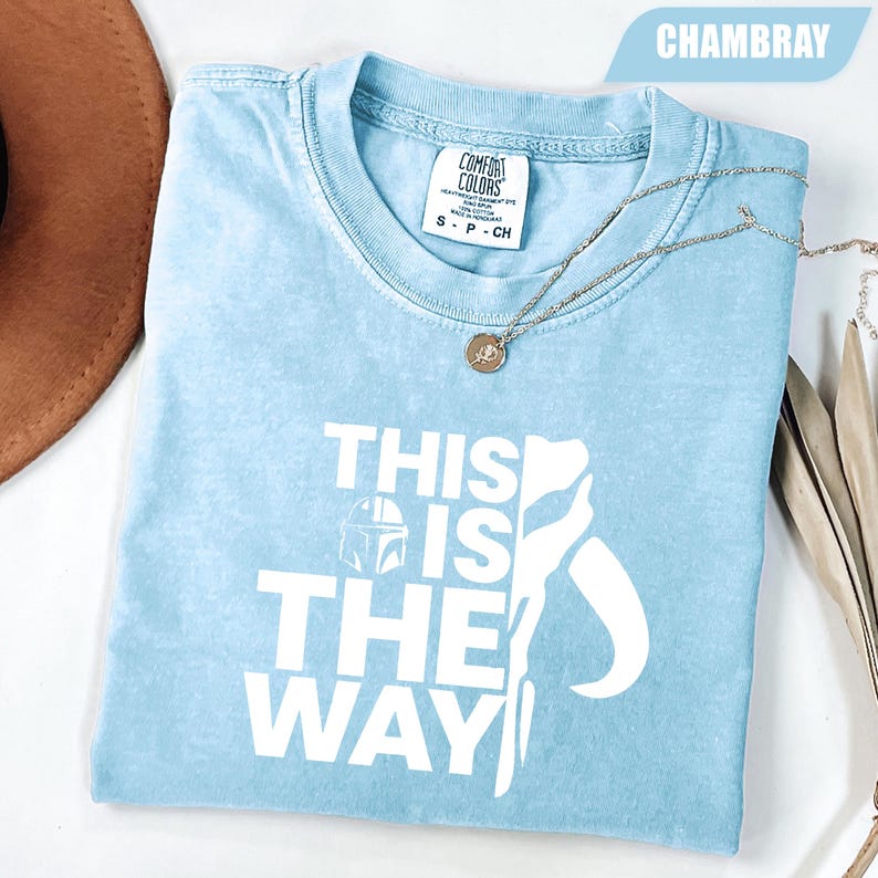 May include: A light blue short-sleeved t-shirt with a white graphic print that reads THIS IS THE WAY, featuring a helmet icon and a stylized silhouette. The shirt has a Comfort Colors brand label at the inner neckline and a crew neck collar.
