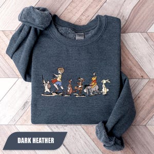 May include: Dark heather gray sweatshirt featuring a cartoon illustration of a boy playing a drum, followed by several animal characters. The text "DARK HEATHER" is displayed at the bottom. The sweatshirt is folded on a light wood grain surface.