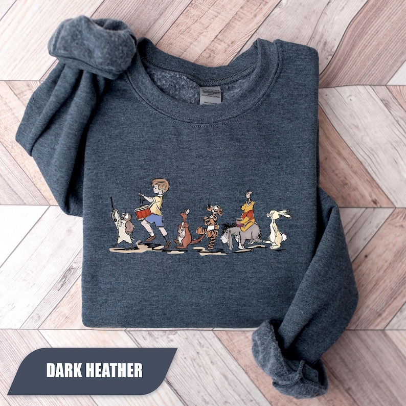 May include: Dark heather gray sweatshirt featuring a cartoon illustration of a boy playing a drum, followed by several animal characters. The text "DARK HEATHER" is displayed at the bottom. The sweatshirt is folded on a light wood grain surface.