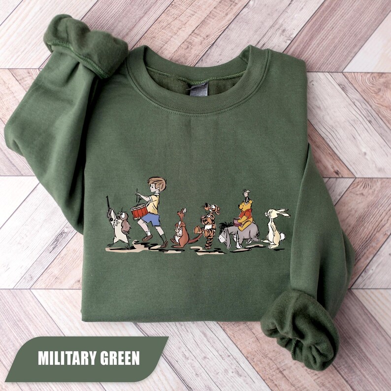 May include: Military green sweatshirt featuring a cartoon illustration of a boy leading a parade of animal characters. The boy is playing a drum, and the animals include a dog, kangaroo, tiger, donkey, bear, and rabbit. The text "MILITARY GREEN" is at the bottom.