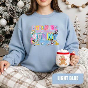 May include: Light blue sweatshirt with the text "I WAS MADE for happy SUNNY days" in colorful letters. The design includes cartoon characters. A white mug with a gingerbread man design is held in the hand.