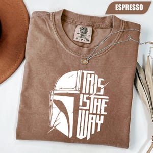 May include: Espresso-colored t-shirt with a white graphic of a helmet and the text "THIS IS THE WAY." The shirt has a crew neck and short sleeves. A silver necklace with a pendant is draped across the shirt.