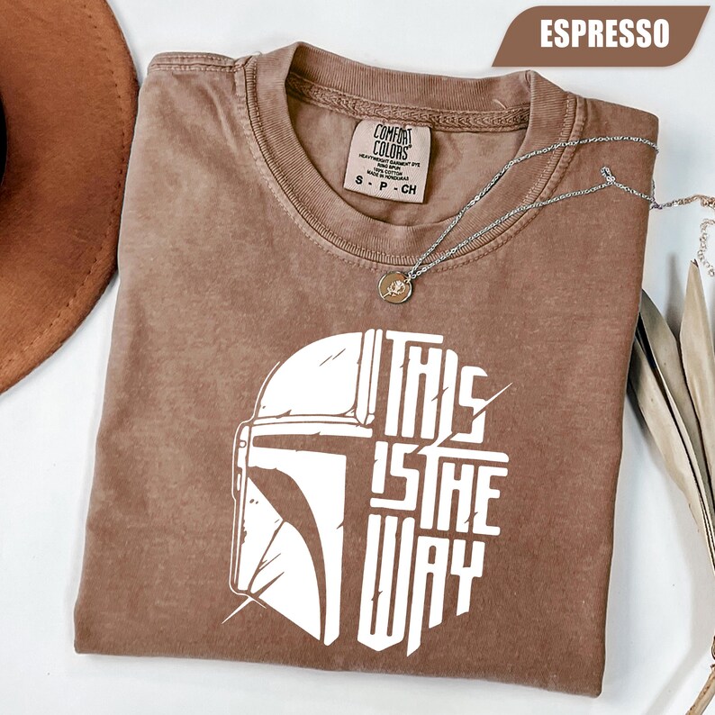 May include: Espresso-colored t-shirt with a white graphic of a helmet and the text "THIS IS THE WAY." The shirt has a crew neck and short sleeves. A silver necklace with a pendant is draped across the shirt.